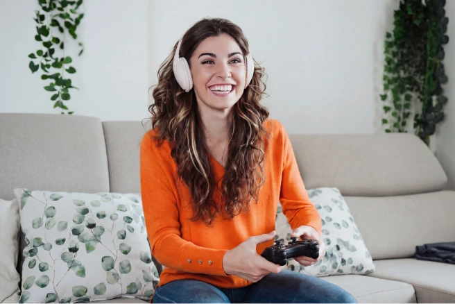 Woman wearing an orange sweater and white headphones, smiling while holding a game controller, seated on a couch.