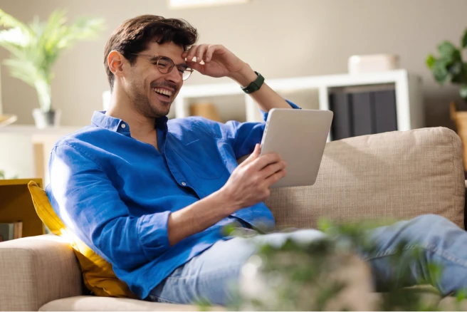 Man in a blue shirt sitting on a couch, smiling while holding a tablet and touching his forehead, in a living room.