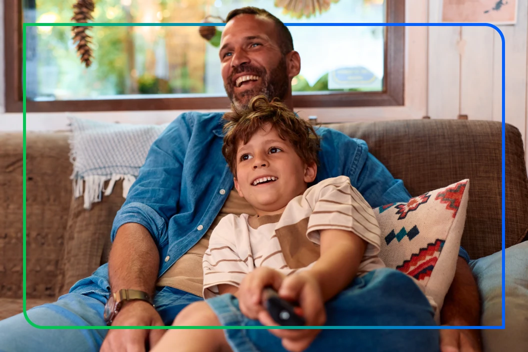 Father and young son laughing together on a couch while the boy holds a TV remote, framed by overlapping blue and green rectangle graphics.
