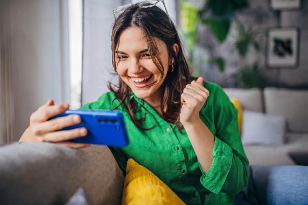 Woman in a green shirt smiling excitedly at a blue smartphone she's holding, making a fist-pump gesture while sitting on a couch with a yellow pillow in a bright living room.
